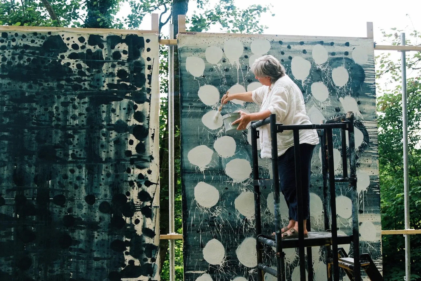 Artist Helen Booth standing on black scaffolding, painting large white polka dots onto dark teal-green canvases in her outdoor studio